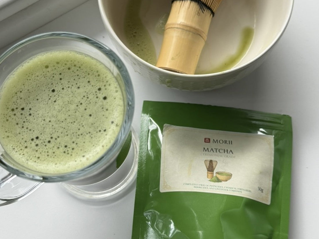 Whisked Morii Uji matcha in a glass, a bowl with a bamboo whisk and the 30 g pack on a windowsill